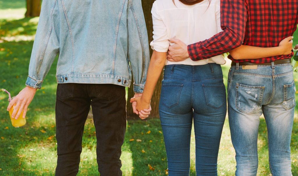 Photo of three people's backs, from their knees to their shoulders. The person on the left is holding hands with the woman in the middle. The woman in the middle also has her hands around the waist of a man on the right.