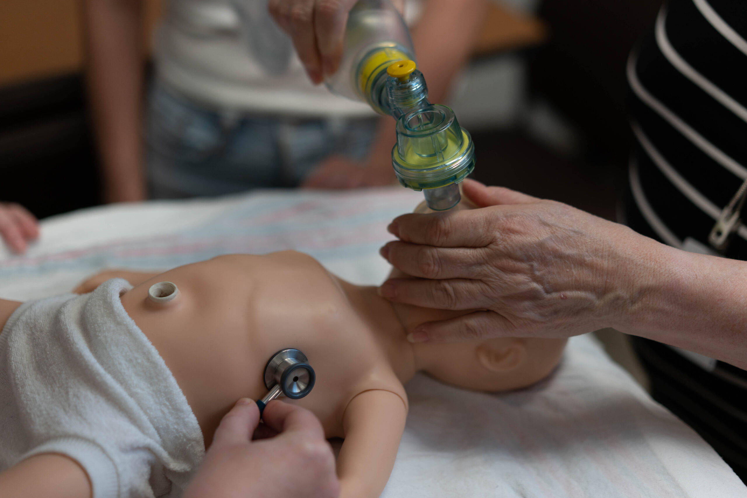 An close-up of an infant manakin being ventilated with a bag valve mask by an instructor while a student auscultates the manakins chest with a stethoscope.
