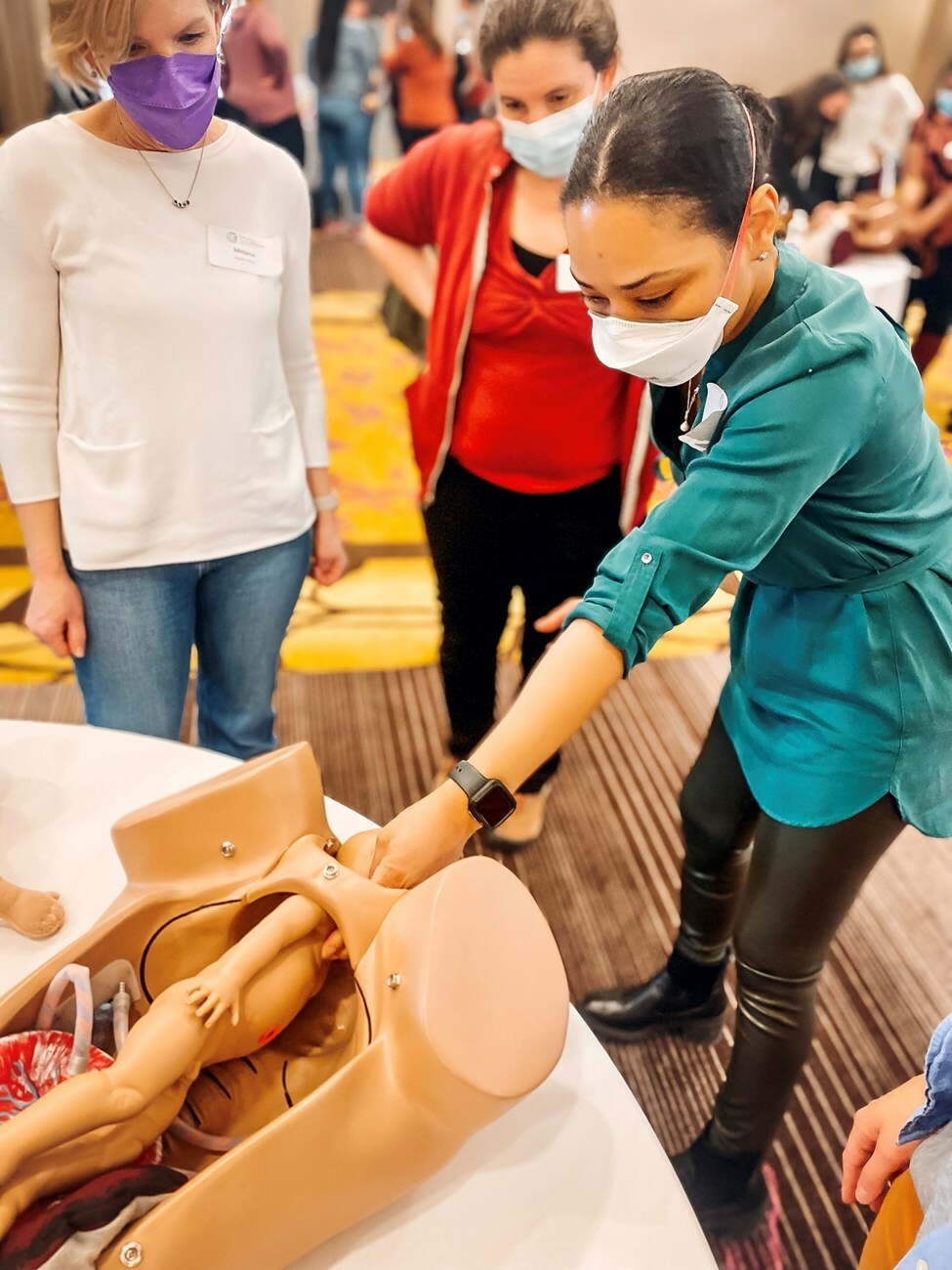 An indoor scene at a training event in a conference area. Three individuals, all wearing face masks, focus on a medical simulation mannequin that replicates a birthing scenario. The woman in the foreground, dressed in a teal shirt, is actively engaged with the mannequin while the other two, one in a white shirt and the other in a red shirt, observe closely. The background shows other participants in groups working on hands-on training.