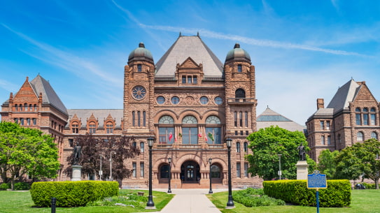 Image of the Ontario Legislative Building, a brownstone structure with Romanesque architectural elements. The building has two rounded towers on either side of the main entrance, with flags displayed above. The front pathway is lined with greenery, and a historical marker stands in the foreground on the grass.