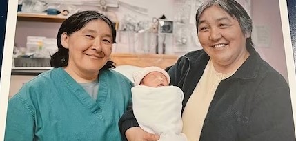 Two smiling women, one in a medical scrubs (Leah Qinuajuak) and the other in a casual jacket (Akinisie Qumaluk), proudly holding a swaddled newborn baby between them in a medical clinic.