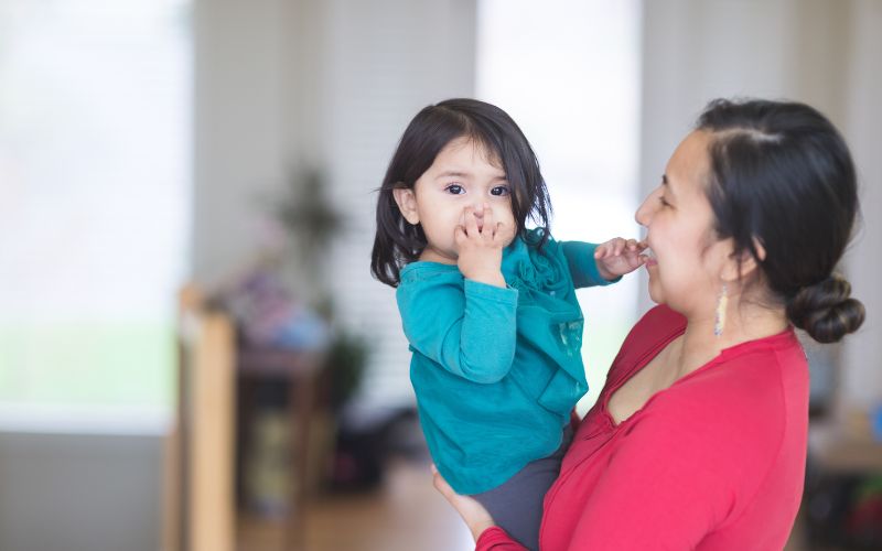 A smiling woman holding a toddler in her arms. The woman is wearing a red top, and the child, who has dark hair, is dressed in a teal-colored shirt. The toddler is looking directly at the camera while sucking their thumb, with their other arm gently resting on the woman's shoulder. The background appears softly blurred, with natural light coming through large windows.