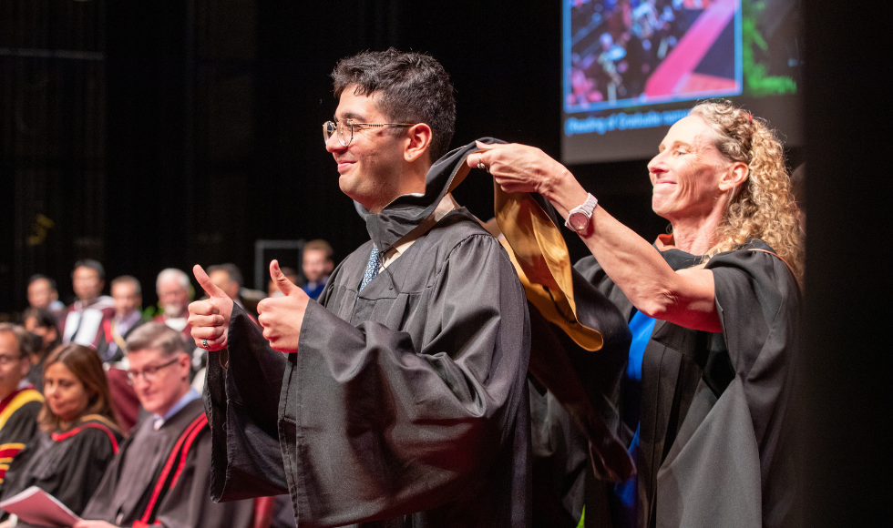 A person in a graduation gown gives a thumbs up while a person standing behind them places a stole over their shoulders.