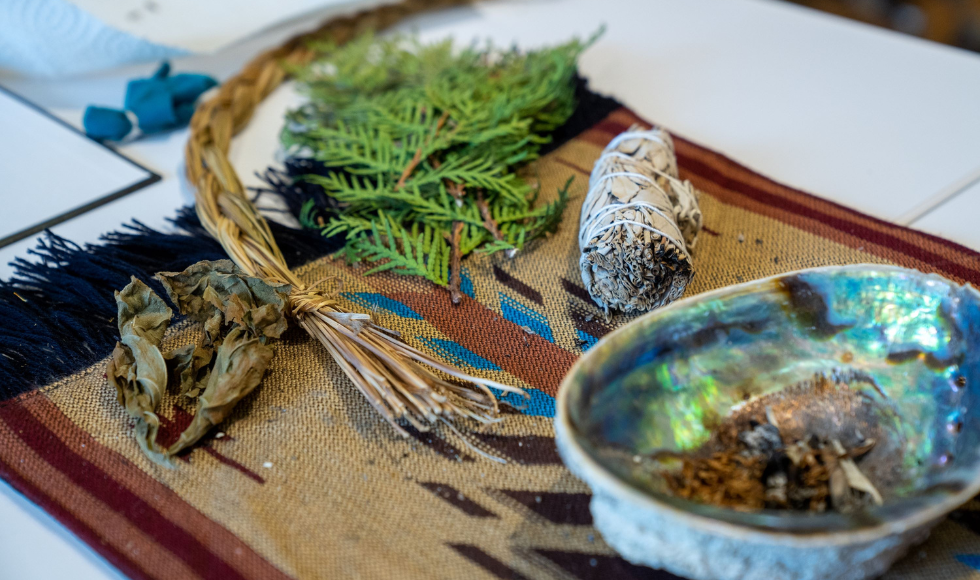 Materials that would be used in a smudging ceremony, including a smudge bowl and some cedar leaves, lie on a table.