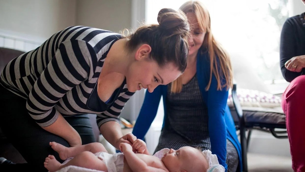 A warm and supportive interaction between a woman and a baby. The woman, wearing a black and white striped shirt, is leaning over and smiling at the baby, who is lying down on a blanket. The baby appears relaxed and comfortable, gazing back at the woman. In the background, two other women are sitting nearby, watching the interaction with smiles, contributing to the overall atmosphere of care, bonding, and support. The setting seems to be a home or a casual, welcoming environment.