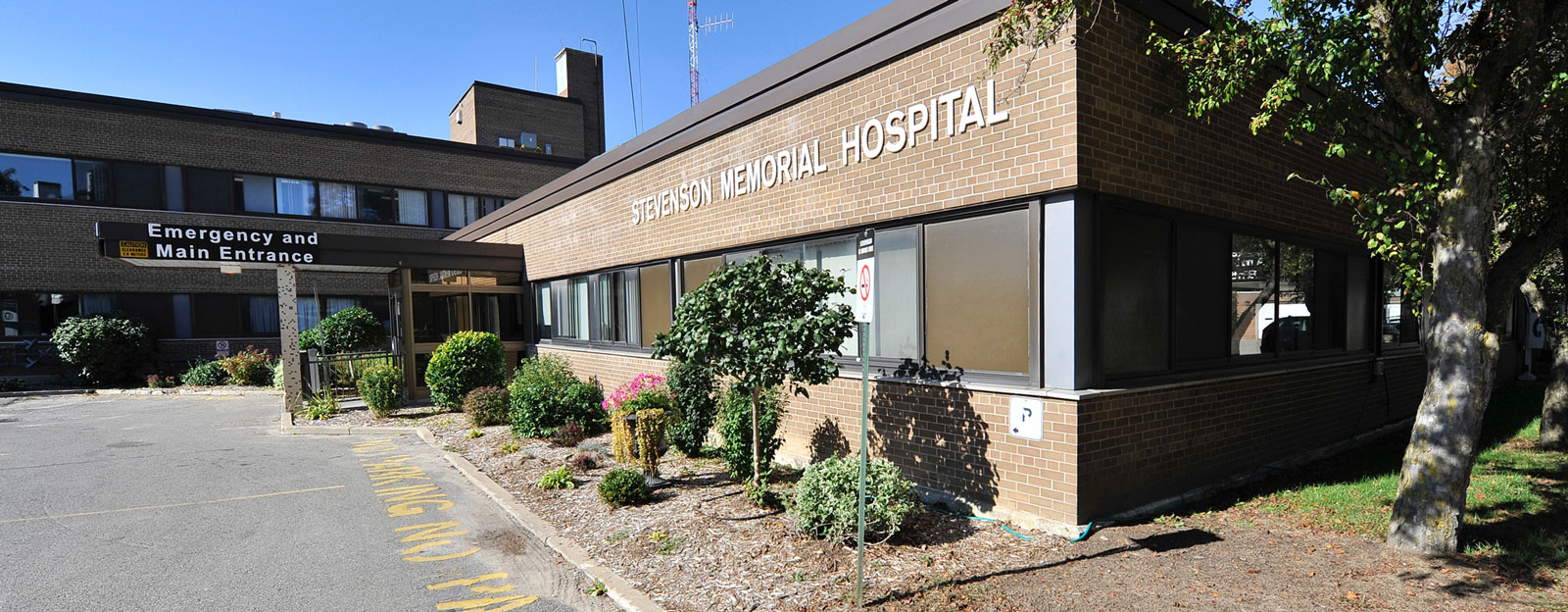 The exterior of Stevenson Memorial Hospital, a brick building with a clean, modern appearance. The hospital's name is prominently displayed on the side of the building in white letters. A well-maintained garden with shrubs and flowers lines the walkway leading to the entrance. The sign for 