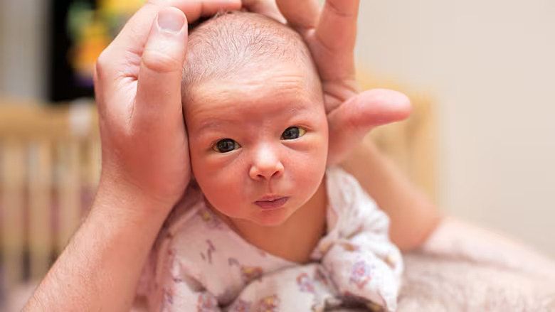 A newborn baby being gently supported by an adult’s hands. The baby, dressed in a light-colored outfit with small patterns, is lying face down and looking directly at the camera with wide, curious eyes.