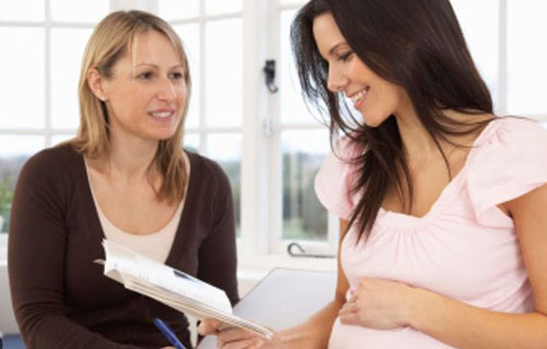 A pregnant woman in a light pink top sitting with a midwife.