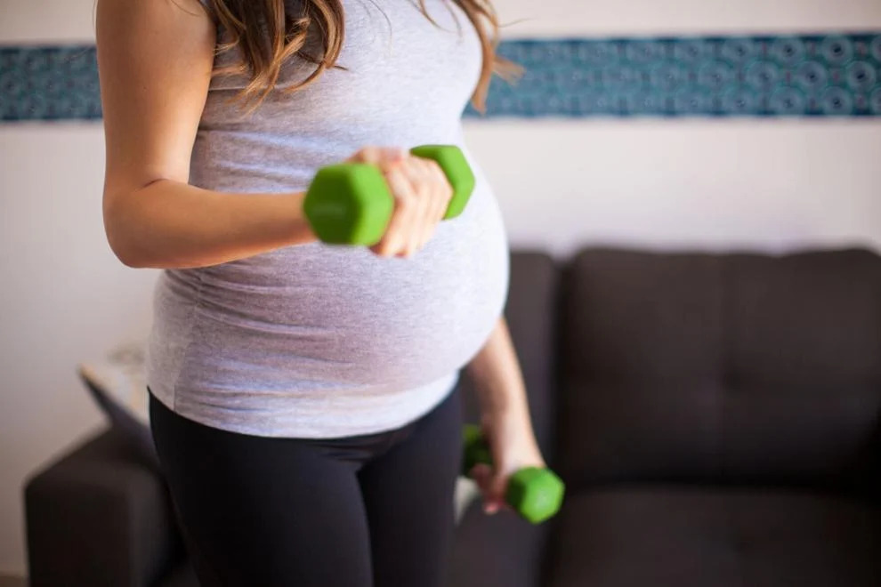 A pregnant woman engaging in light exercise. She is holding two small green dumbbells, lifting one in each hand, while wearing a fitted grey tank top and black leggings that accentuate her baby bump.